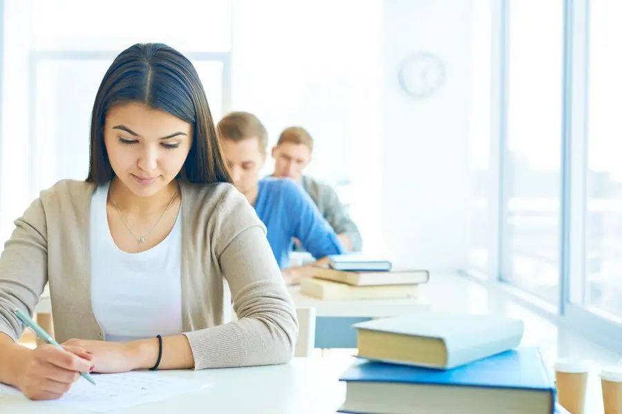 A young woman is writing an exam in a bright classroom with books on the desk